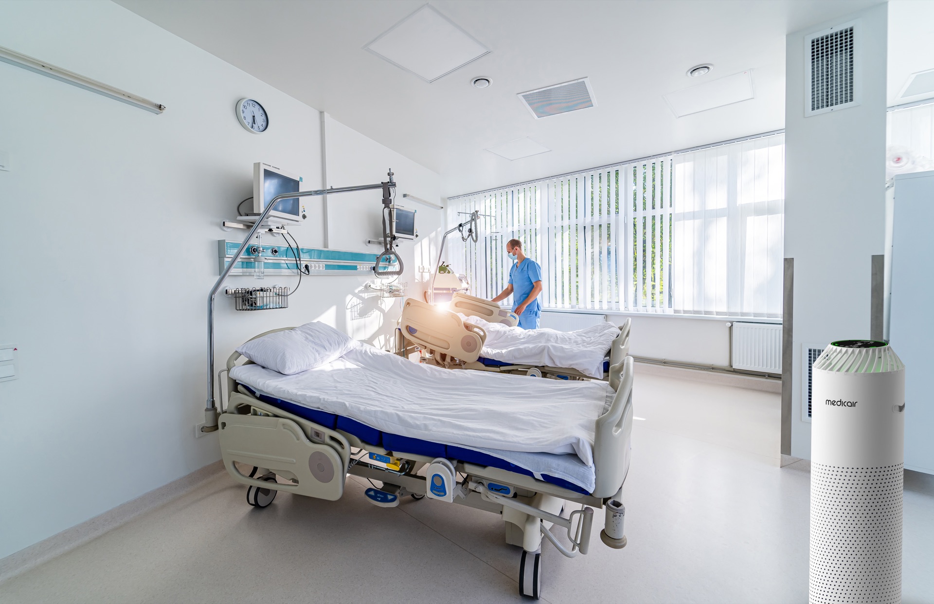 MedicAir Pro air purifier beside two beds in a bright hospital ward with a clinician in blue scrubs