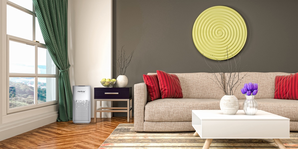 MedicAir Pro Mini air purifier by a window next to a side table in a living room with a beige sofa and red cushions
